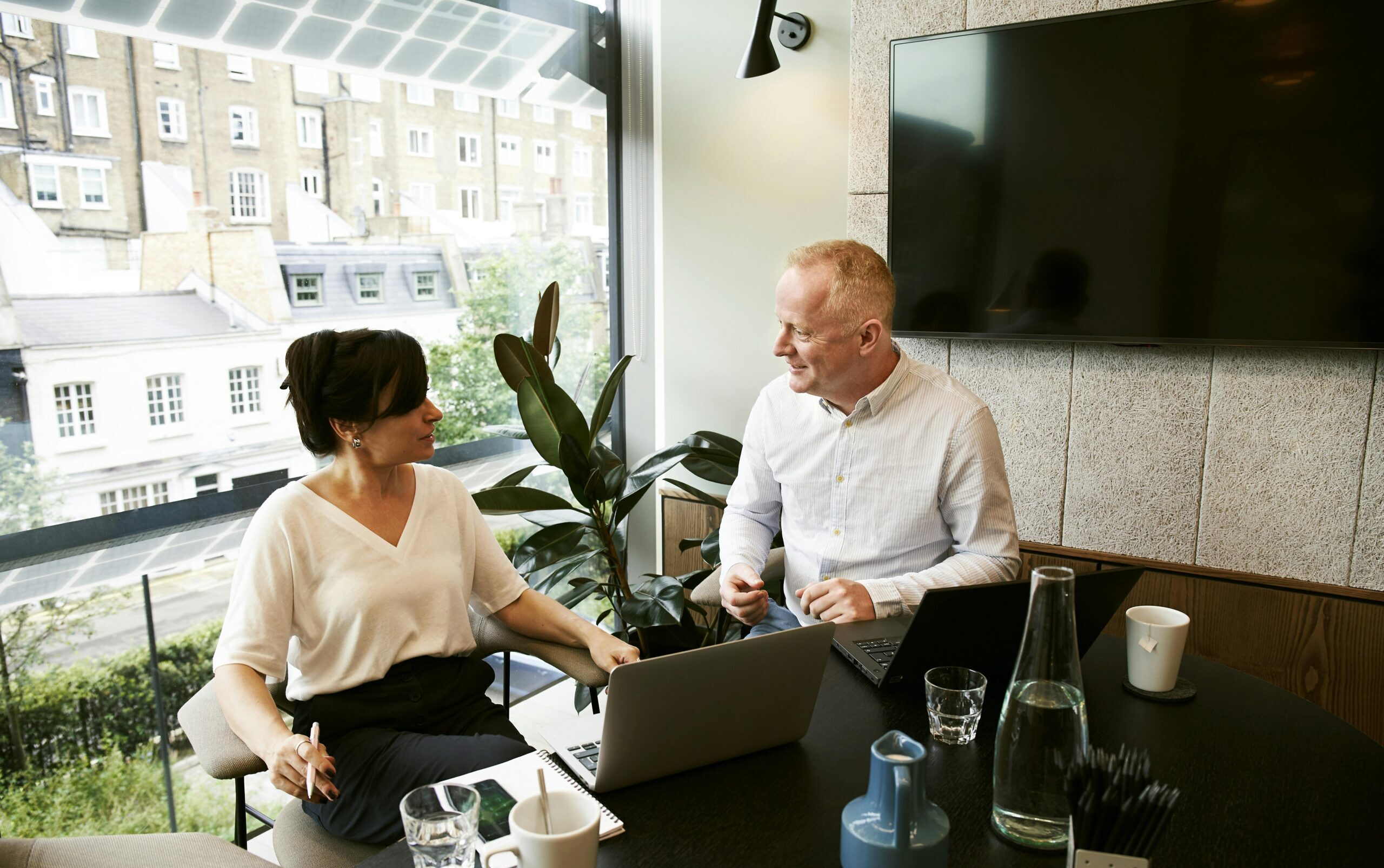 Two business professionals engaging in a team meeting in a modern London office with a large window view.